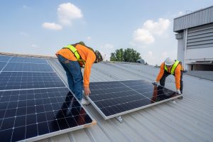 Two workers in safety gear install solar panels on a metal rooftop under a clear sky, showcasing expert Solar Panel Installation in East Baton Rouge Parish.