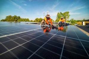 Two workers in safety gear install solar panels on a rooftop under a clear blue sky, showcasing Solar Installation Services St. Charles Parish, with trees and buildings visible in the background.