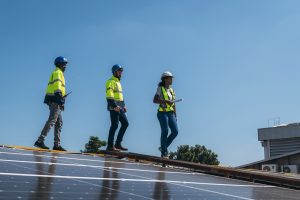 Three people in safety gear walk on a rooftop in Lafourche Parish, carrying clipboards and inspecting the solar panel installation under a clear sky.
