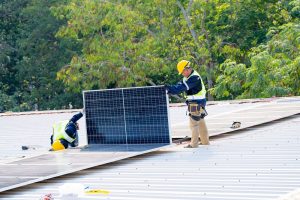 Two workers in safety gear install a large solar panel on a metal rooftop, surrounded by greenery—exemplifying the quality of Solar Installation Services St. Charles Parish has to offer.