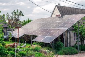 A house with a pitched roof is partially covered by a large array of St John the Baptist Parish Solar Panels installed on metal supports in a garden area filled with greenery and garden ornaments.
