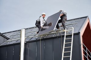 Two workers wearing safety gear, representing top Solar Installers in Tangipahoa Parish, install a solar panel on the slanted roof of a building, with a ladder and metal chimney visible.