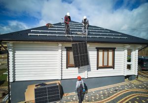 Solar Installers in Plaquemines Parish work together as three workers install solar panels on the roof of a white house—two on the roof and one on the ground assisting with lifting a panel.